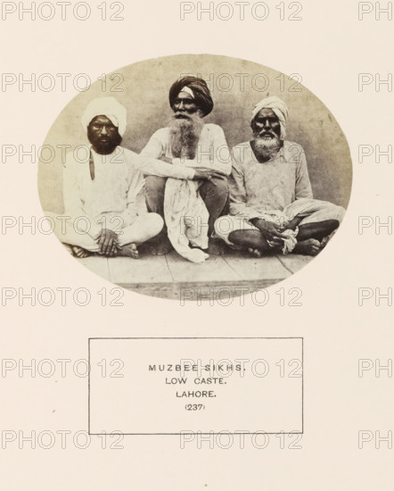 Portrait of three unknown men from the Mazhabi Sikh community of Lahore, Punjab, Pakistan, photograph, page, number: '(237)', inscription: 'MUZBEE SIKHS., LOW CASTE., LAHORE.', Lahore, c. 1862 - in or before 1872, photographer, paper, albumen print, height 95 mm x width 120 mm, human races, peoples, nationalities, Three seated men appear inside a circular frame, each wearing light-colored loose clothing and turbans, with visible facial hair and bare feet; the man in the center has a long beard and folded arms resting on his knees, the two men on either side sit cross-legged with their hands resting on their legs, and a simple floorboard surface is visible beneath them.