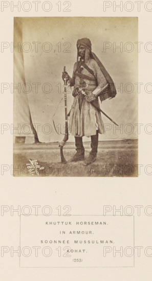 Portrait of a horseman with a musket and saber from the Khattak tribe of Kohat, Khyber Pakhtunkhwa, Pakistan, photograph, page, number: '(253)', inscription: 'KHUTTUK HORSEMAN., IN ARMOUR., SOONNEE MUSSULMAN., KOHAT.', Kohat, 1861, photographer, paper, albumen print, height 110 mm x width 86 mm, riding a horse, ass, or mule, rider, horseman, firearms: rifle, human races, peoples, nationalities, hacking and thrusting weapons (with NAME), A full-length sepia photograph of a person standing on a textured floor against a cloth backdrop, wearing layered garments including a draped head covering and a long tunic with a belt, holding a long firearm upright with one hand and resting the other hand near a sheathed sword at the waist, with boots visible and a strap or sash crossing the chest, and a small signature or marking near the lower left of the photo.