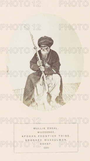Portrait of Mullik Ensal, a man with a rifle from the Wazir tribe of Kohat, Khyber Pakhtunkhwa, Pakistan, photograph, page, The Wazir tribe is also known as Waziri., number: '(254)', inscription: 'MULLIK ENSAL., WUZEEREE., AFGHAN FRONTIER TRIBE., SOONNEE MUSSULMAN., KOHAT.', Kohat, c. 1862 - in or before 1872, photographer, paper, albumen print, height 120 mm x width 95 mm, head-gear: turban, firearms: rifle, human races, peoples, nationalities, An oval-framed sepia photograph shows an older man seated on a striped surface, wearing a dark turban and a dark long coat over lighter loose trousers, with a long white beard and a solemn facial expression, holding a long decorated rifle vertically across his body with both hands, his bare feet visible beneath his clothing.