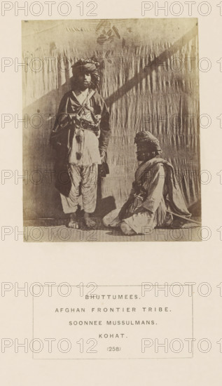 Portrait of two unknown tribesmen with turbans from Kohat, Khyber Pakhtunkhwa, Pakistan, photograph, page, number: '(258)', inscription: 'BHUTTUMEES., AFGHAN FRONTIER TRIBE., SOONNEE MUSSULMANS., KOHAT.', Kohat, c. 1862 - in or before 1872, photographer, paper, albumen print, height 109 mm x width 88 mm, head-gear: turban, human races, peoples, nationalities, A sepia-toned photograph showing two men in traditional clothing against a draped backdrop, one man standing barefoot with layered garments and a turban, the other seated on the floor wearing similar attire and a turban, both facing forward, with the standing man's shadow cast on the backdrop and a rectangular caption box below the photograph.