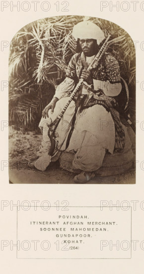 Portrait of an unknown nomadic trader from the Powindah tribe of Kohat, Khyber-Pakhtunkhwa, Pakistan, photograph, page, The Powindah tribe is also known as Kochi., number: '(264)', inscription: 'POVINDAH., ITINERANT AFGHAN MERCHANT., SOONNEE MAHOMEDAN., GUNDAPOOR.', Kohat, 1861, photographer, paper, albumen print, height 115 mm x width 87 mm, head-gear: turban, firearms: rifle, human races, peoples, nationalities, A seated person wearing a wrapped head covering and layered garments sits on a round cushion in front of tall leafy plants, holding a long decorated rifle across their body; the clothing includes a patterned outer garment over lighter sleeves and loose trousers, and the person has dark hair visible under the head covering and a neutral facial expression while looking slightly off camera.
