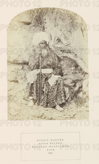 Portrait of Khaddir Bukhsh, a landholder in Sindh, Pakistan, photograph, page, inscription: 'KHADIR BUKHSH., KHOSA BELOCH., SOONNEE MUSSULMAN., SIND.', number: '292.', photographer: Henry Charles Baskerville Tanner (possibly), Sindh, c. 1862 - in or before 1872, paper, albumen print, height 148 mm x width 107 mm, head-gear: turban, human races, peoples, nationalities, A sepia photograph shows an older man seated against a textured rocky background with roots and rough surfaces. He wears a patterned long robe and a large wrapped turban, and he has a long beard and mustache. He holds a long staff with one hand and rests the other hand on his lap, where a light-colored cloth is draped. His shoes are visible beneath the hem of his robe and the ground around him is covered with small stones and debris. The image is framed with rounded top corners and surrounded by a wide light border.