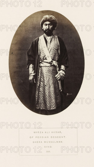 Portrait of Mirza Ali Akbar, a nobleman of Persian descent, Sindh, Pakistan, photograph, page, inscription: 'MIRZA ALI AKBAR, OF PERSIAN DESCENT., SHEEA MUSSULMAN., SIND.', number: '295.', photographer: Henry Charles Baskerville Tanner (possibly), c. 1862 - in or before 1872, paper, albumen print, height 139 mm x width 100 mm, head-gear: turban, prince, human races, peoples, nationalities, An oval sepia photograph shows a standing man facing forward wearing a patterned long robe with a sash and a darker outer coat, a turban on his head, and a mustache and beard; his left hand rests near a sheathed sword at his waist while his right arm hangs by his side, and the oval image is centered on a light background with a rectangular bordered area below containing printed lines.