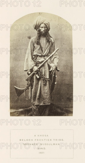 Portrait of an unknown man with a rifle from the Khosa Baloch tribe in Sindh, Pakistan, photograph, page, inscription: 'A KHOSA., BELOCH FRONTIER TRIBE., SOONNEE MUSSULMAN., SIND.', number: '(307)', photographer: Henry Charles Baskerville Tanner (possibly), Sindh, photographer: William Robert Houghton (possibly), c. 1862 - in or before 1872, paper, albumen print, height 129 mm x width 79 mm, head-gear: turban, firearms: rifle, human races, peoples, nationalities, A sepia-toned portrait of a standing man wearing a wrapped turban and long robe, with long hair and a beard, holding a long staff diagonally across his body and carrying a curved rifle and other objects at his waist, set inside an arched vignette on a pale background.