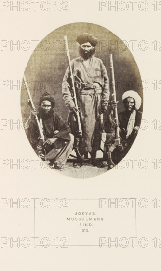 Portrait of three unknown men from the Jokhio Sindhi tribe in Sindh, Pakistan, photograph, page, inscription: 'JOKYAS., MUSSULMANS., SIND.', number: '313.', photographer: Henry Charles Baskerville Tanner (possibly), Sindh, photographer: William Robert Houghton (possibly), c. 1862 - in or before 1872, paper, albumen print, height 120 mm x width 95 mm, head-gear: turban, firearms: rifle, human races, peoples, nationalities, A vignette-style oval photograph centered on a pale background showing three dark-skinned men posed with long rifles; one man stands in the middle wearing a turban and loose robe with a belt, his hands resting on the rifles at his sides, while the other two men are seated or crouched on either side holding their rifles upright, each wearing head coverings and loose garments, with one wearing a wrapped turban and the other a cap; the image has a sepia tone and visible grain and the oval photograph sits above a rectangular blank area on the page.