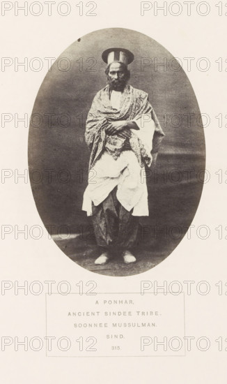 Portrait of an unknown man from the Punar Sindhi tribe in Sindh, Pakistan, photograph, page, inscription: 'A PONHAR., ANCIENT SINDEE TRIBE., SOONNEE MUSSULMAN., SIND.', number: '315.', photographer: Henry Charles Baskerville Tanner (possibly), Sindh, photographer: William Robert Houghton (possibly), c. 1862 - in or before 1872, paper, albumen print, height 139 mm x width 100 mm, head-gear: hat, human races, peoples, nationalities, A sepia-toned oval photograph centered on a pale background shows a standing man facing forward wearing layered traditional clothing and a tall rounded hat; a patterned shawl is draped over his shoulders and crossed arms, a lighter garment hangs around his waist over striped trousers, and he wears light shoes.