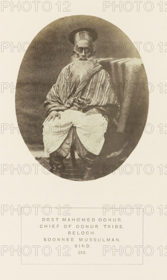 Portrait of Dost Mahomed, a landholder and chieftain from the Baloch community in Sindh, Pakistan, photograph, page, number: '316.', inscription: 'DOST MAHOMED OONUR., CHIEF OF OONUR TRIBE., BELOCH., SOONNEE MUSSULMAN., SIND.', photographer: Henry Charles Baskerville Tanner (possibly), Sindh, photographer: William Robert Houghton (possibly), c. 1862 - in or before 1872, paper, albumen print, height 106 mm x width 86 mm, head-gear: hat, ruler, sovereign, human races, peoples, nationalities, An oval sepia-toned photograph of an elderly man seated facing forward, wearing a tall rounded hat and a long striped robe draped over his shoulders, with a long white beard and hands resting on his knees, light-colored loose trousers gathered over darker footwear, and a textured cloth draped over the back of the chair beside him, all set against a neutral background and mounted on a pale sheet.