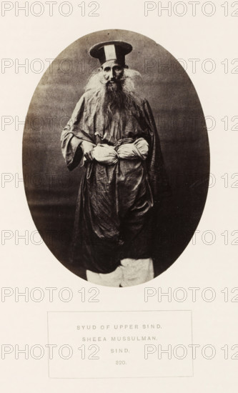 Portrait of an unknown landholder in Sindh, Pakistan, photograph, page, inscription: 'SYUD OF UPPER SIND., SHEEA MUSSULMAN., SIND.', number: '320.', photographer: Henry Charles Baskerville Tanner (possibly), Sindh, photographer: William Robert Houghton (possibly), c. 1862 - in or before 1872, paper, albumen print, height 139 mm x width 101 mm, head-gear: hat, human races, peoples, nationalities, An oval-framed black-and-white photograph shows an elderly man standing and facing forward wearing a tall round hat with a contrasting vertical band, a long flowing robe with wide sleeves, and loose trousers; he has a long full beard and wispy hair at his temples, visible facial features with a solemn expression, and he holds a wrapped bundle at his waist with both hands.