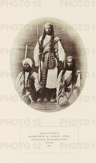 Portrait of three unknown men with weapons from the Brahui community of Kalat, Balochistan, Pakistan, photograph, page, number: '321.', inscription: 'BRAHOOEES., INHABITANTS OF KHELAT STATE., SOONNEE MAHOMEDANS., SIND.', photographer: Henry Charles Baskerville Tanner (possibly), photographer: William Robert Houghton (possibly), c. 1862 - in or before 1872, paper, albumen print, height 128 mm x width 105 mm, weapons, human races, peoples, nationalities, An oval sepia photograph shows three men wearing traditional garments and head coverings, two seated at the sides and one standing in the center; the standing man has long hair or braids and a draped shawl over one shoulder, and each man holds a long firearm with visible stocks and barrels; the seated men wear turbans or headwraps and have long hair and beards, with layered clothing and sashes, and a patterned textile is visible beneath their feet; the photograph is set against a neutral backdrop and framed by a wide light border with a ...