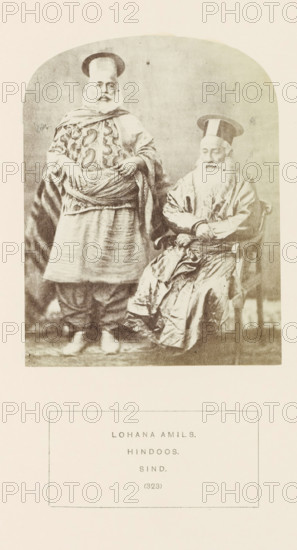 Portrait of two unknown men from the Lohana Amil community in Sindh, Pakistan, photograph, page, number: '(323)', inscription: 'LOHANA AMILS., HINDOOS., SIND.', photographer: Henry Charles Baskerville Tanner (possibly), Sindh, photographer: William Robert Houghton (possibly), c. 1862 - in or before 1872, paper, albumen print, height 123 mm x width 96 mm, head-gear: hat, folk costume, regional costume, human races, peoples, nationalities, A sepia-toned photograph shows two older men wearing traditional garments and tall flat hats; one man stands with hands resting on a wrapped sash around his waist, wearing a patterned tunic and layered skirt-like lower garment with loose trousers beneath, while the other man sits in a wooden armchair with hands folded, wearing a long shiny robe with many folds and a long white beard; the background is plain and the image has rounded top corners and a light border around it.
