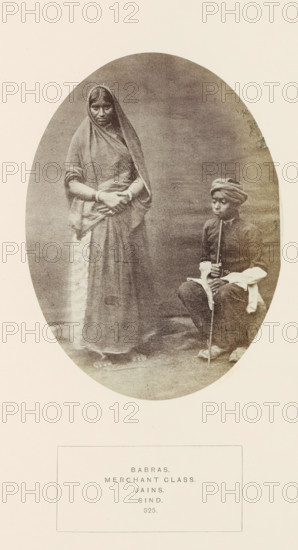 Portrait of an unknown woman and her son from the Bhabra community in Sindh, Pakistan, photograph, page, inscription: 'BABRAS., MERCHANT CLASS., JAINS., SIND.', number: '325.', photographer: Henry Charles Baskerville Tanner (possibly), Sindh, photographer: William Robert Houghton (possibly), c. 1862 - in or before 1872, paper, albumen print, height 146 mm x width 109 mm, head-gear: turban, human races, peoples, nationalities, A sepia-toned oval photograph shows two figures against a plain backdrop. On the left stands a woman draped in layered fabric that covers her head and body, her hands clasped at waist level and feet visible beneath the hem. On the right a seated youth wears a turban and a long-sleeved upper garment with contrasting lighter fabric at the cuffs, holding a long slender staff vertically with both hands and looking slightly downward. The floor beneath them appears textured and the overall image has a soft, aged photographic quality.