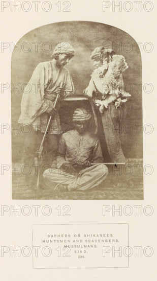 Portrait of three unknown hunters from Sindh, Pakistan, with a dead bird and rifles, photograph, page, number: '326.', inscription: 'DAPHERS OR SHIKAREES., HUNTSMEN AND SCAVENGERS., MUSSULMANS., SIND.', photographer: Henry Charles Baskerville Tanner (possibly), Sindh, photographer: William Robert Houghton (possibly), c. 1862 - in or before 1872, paper, albumen print, height 146 mm x width 108 mm, head-gear: turban, hunter, firearms: rifle, human races, peoples, nationalities, A sepia-toned photograph framed with an arched top shows three men wearing turbans and loose traditional garments; one man stands on the left leaning on a long rifle with both hands, his head tilted downward, another man stands on the right facing slightly toward the center wearing a textured fur or feathered wrap over his shoulders and resting an elbow on a horizontal surface, and a third man sits cross-legged on the ground in front of them holding a long firearm across his lap and looking down; the background is...