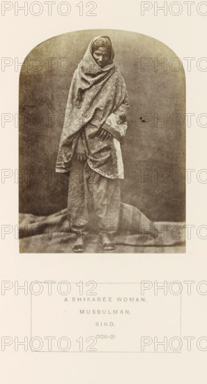Portrait of an unknown huntress with a headscarf from Sindh, Pakistan, photograph, page, number: '(326-2)', inscription: 'A SHIKAREE WOMAN., MUSSULMAN., SIND.', photographer: Henry Charles Baskerville Tanner (possibly), Sindh, photographer: William Robert Houghton (possibly), c. 1862 - in or before 1872, paper, albumen print, height 116 mm x width 87 mm, head-gear (with NAME), human races, peoples, nationalities, A sepia-toned photograph shows a standing woman draped in a patterned shawl that covers her head and wraps around her body, leaving only part of her face and hands visible; she wears loose trousers and flat footwear, and stands on a textured cloth or backdrop with a plain, slightly mottled background behind her.