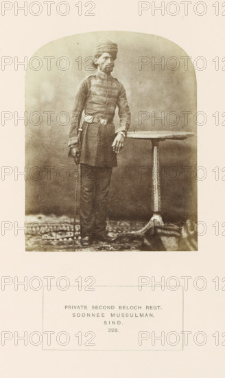 Portrait of a soldier in the second Baloch regiment, Sindh, Pakistan, photograph, page, inscription: 'PRIVATE SECOND BELOCH REGT., SOONNEE MUSSULMAN., SIND.', number: '328.', photographer: Henry Charles Baskerville Tanner (possibly), photographer: William Robert Houghton (possibly), c. 1862 - in or before 1872, paper, albumen print, height 123 mm x width 96 mm, the soldier, the soldier's life, human races, peoples, nationalities, A sepia-toned portrait of a standing man wearing a turban and a long, buttoned tunic with horizontal trim across the chest, a belt with a buckle, straight trousers, and shoes; he rests one hand on a round table with an ornate central pedestal and holds a thin cane in his other hand, standing on a patterned rug against a plain backdrop.