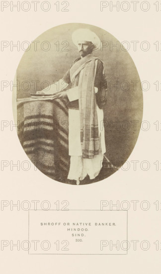 Portrait of an unknown money changer in Sindh, Pakistan, photograph, page, inscription: 'SHROFF OR NATIVE BANKER., HINDOO., SIND.', number: '330.', photographer: Henry Charles Baskerville Tanner (possibly), Sindh, photographer: William Robert Houghton (possibly), c. 1862 - in or before 1872, paper, albumen print, height 129 mm x width 104 mm, head-gear: turban, banker, human races, peoples, nationalities, An oval sepia photograph shows a man standing beside a small table draped with a striped blanket; he wears a wrapped turban and layered garments including a long outer shawl draped over one shoulder and a light-colored lower garment, and his hands rest on a stack of flat objects on the table while his face looks toward the camera.