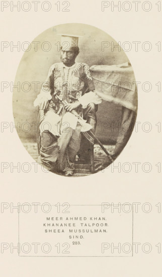 Portrait of Mir Ahmed Khan, Sindh, Pakistan, photograph, page, inscription: 'MEER AHMED KHAN., KHANANEE TALPOOR., SHEEA MUSSULMAN., SIND.', number: '283.', photographer: Henry Charles Baskerville Tanner (possibly), c. 1862 - in or before 1872, paper, albumen print, height 107 mm x width 87 mm, ruler, sovereign, human races, peoples, nationalities, An oval sepia photograph shows a seated man wearing a patterned long coat with decorative embroidery and wide cuffs, striped loose trousers, and a flat square hat; he sits beside a draped table with one hand resting on his knee and the other holding an object, and a sword or long stick leans against the chair extending toward the floor.