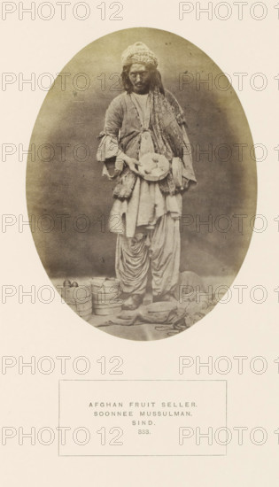 Portrait of an unknown Afghan fruit seller in Sindh, Pakistan, photograph, page, inscription: 'AFGHAN FRUIT SELLER., SOONNEE MUSSULMAN., SIND.', number: '333.', photographer: Henry Charles Baskerville Tanner (possibly), Sindh, photographer: William Robert Houghton (possibly), c. 1862 - in or before 1872, paper, albumen print, height 147 mm x width 108 mm, merchant, salesman, human races, peoples, nationalities, An oval-framed sepia photograph of a standing person wearing layered loose clothing and a patterned headwrap, holding a shallow round tray with round items; the clothing includes a long draped garment, a sash, and pleated lower trousers, with visible worn shoes; at the subject's feet are stacked round containers and scattered cloth bundles and small objects; the background is plain and darker within the oval while the surrounding border is light.