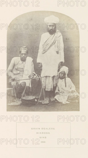 Portrait of three unknown grain dealers in Sindh, Pakistan, photograph, page, inscription: 'GRAIN DEALERS., HINDOOS., SIND.', number: '(334)', photographer: Henry Charles Baskerville Tanner (possibly), Sindh, photographer: William Robert Houghton (possibly), c. 1862 - in or before 1872, paper, albumen print, height 144 mm x width 105 mm, merchant, salesman, human races, peoples, nationalities, A faded monochrome photograph shows three people positioned against a plain backdrop inside an arched frame. One person is seated on the left, wearing a light long-sleeved garment with a cloth draped over one shoulder and a cap on the head, holding a horizontal pole balanced on their knee with suspended pans. A tall standing person in the center wears a long light robe and a wrapped head covering, and a patterned shawl draped across one shoulder, with one hand resting near the pole. A smaller seated person on the right wears a light tunic and a turban-like head wrap, sitting cross-legged with han...