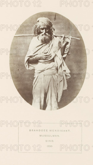 Portrait of an unknown mendicant from the Brahui community in Sindh, Pakistan, photograph, page, inscription: 'BRAHOOEE MENDICANT., MUSSULMAN., SIND.', number: '(336)', photographer: Henry Charles Baskerville Tanner (possibly), Sindh, photographer: William Robert Houghton (possibly), c. 1862 - in or before 1872, paper, albumen print, height 120 mm x width 95 mm, religious functions, Hinduism, Buddhism, Jainism, human races, peoples, nationalities, An oval sepia photograph shows an older bearded man standing and facing forward, wearing layered loose cloth draped around his body and a turban on his head, with one hand resting on his chest and the other holding a long thin pole balanced across his shoulders; the background inside the oval is plain and slightly mottled, and the photograph is centered on a pale rectangular page with blank space surrounding it.