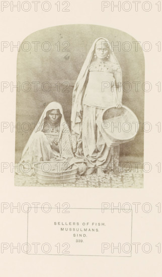 Portrait of two unknown fish sellers in Sindh, Pakistan, photograph, page, inscription: 'SELLERS OF FISH., MUSSULMANS., SIND.', number: '339.', photographer: Henry Charles Baskerville Tanner (possibly), Sindh, photographer: William Robert Houghton (possibly), c. 1862 - in or before 1872, paper, albumen print, height 123 mm x width 97 mm, merchant, salesman, head-gear (with NAME), human races, peoples, nationalities, A faded sepia photograph shows two women in traditional draped garments and head coverings within an arched frame, one woman seated on a patterned floor holding a shallow basket in front of her and the other standing beside her holding a larger round sieve or shallow basket at her side; both wear layered skirts and shawls, and the standing woman has visible earrings.