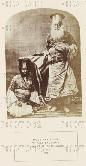 Portrait of Ali Khan and an attendent Sindh, Pakistan, photograph, page, inscription: 'DOST ALI KHAN., THORA TALPOOR., SHEEA MUSSULMAN., SIND.', number: '285.', photographer: Henry Charles Baskerville Tanner (possibly), Sindh, c. 1862 - in or before 1872, paper, albumen print, height 139 mm x width 96 mm, ruler, sovereign, human races, peoples, nationalities, A sepia-toned portrait shows two men in traditional clothing against a plain backdrop, one seated on the floor wearing layered garments with a patterned upper piece and a wrapped sash, a small flat-topped headpiece, and a calm facial expression, the other standing with a long white beard, wearing a long robe and a tall cylindrical hat, holding a wrapped bundle and draped fabric, with a blanket or cloth hanging between them and a patterned rug beneath.