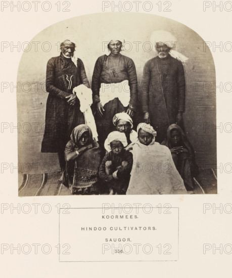 Portrait of eight people and children from the Kurmi community of Sagar, Madhya Pradesh, India, photograph, page, inscription: 'KOORMEES., HINDOO CULTIVATORS., SAUGOR.', number: '356.', photographer: James John Waterhouse (possibly), Sagar, 1862, Central India, paper, albumen print, height 118 mm x width 129 mm, human races, peoples, nationalities, A sepia photograph shows a group portrait of people arranged in two rows against a plain curved backdrop, with three adults standing in the back and several individuals seated or crouched in front; the standing figures wear long dark garments and headwraps, one holds a light cloth, and the central standing person has a contrasting light cloth around the waist, while the seated group features patterned shawls, cloths wrapped around heads, and a large light blanket draped over one person, all on a striped floor covering.