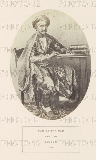 Portrait of Rao Krishn Rao, a pundit from Sagar at a reading table, Madhya Pradesh, India, photograph, page, inscription: 'RAO KRISHN RAO., HINDOO., SAUGOR.', number: '357.', photographer: James John Waterhouse (possibly), Sagar, 1862, Central India, paper, albumen print, height 148 mm x width 109 mm, scholar in his study, human races, peoples, nationalities, An oval-framed black-and-white portrait of an older man seated beside a table, wearing a turban and layered patterned robes with a draped shawl over one shoulder, his hands resting on the table and a stack of books beside him, a carved wooden chair partly visible behind him, a patterned floor covering beneath his feet, and a rectangular border containing printed lines below the oval.