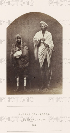 Portrait of a soldier Nar Sing and his wife Chundi from the Bhil tribe of Jhabua, Madhya Pradesh, India, photograph, page, inscription: 'BHEELS OF JHABOOA., CENTRAL INDIA.', number: '365.', photographer: James John Waterhouse (possibly), Jhabua, 1862, Central India, paper, albumen print, height 115 mm x width 79 mm, human races, peoples, nationalities, A sepia photograph framed by an arched top shows a standing man and a seated or slightly crouched person beside him against a plain backdrop; the man is draped in a wrapped cloth over one shoulder and wears a turban, holding a long curved object and another stick-like item across his body, while the person next to him is wrapped in patterned fabric with a hood covering the head and cradling a bundle, and both figures have bare feet with decorative bands visible on the lower legs of the seated person.