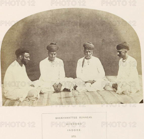 Portrait of four unknown merchants originally from Shekhawati, Rajasthan, based in Indore, Madhya Pradesh, India, photograph, page, number: '372.', inscription: 'SHEKHAWATTEE BUNNEAS., HINDOOS., INDORE.', photographer: James John Waterhouse (possibly), Indore, 1862, Central India, paper, albumen print, height 115 mm x width 168 mm, head-gear: turban, human races, peoples, nationalities, A sepia photograph shows four men seated on a striped cloth floor against a plain curved backdrop, all wearing light-colored tunics and headwraps, with two men facing inward toward the center and the two outer men turned slightly inward; their hands rest on their laps or knees and one man wears a pendant at his chest, while a folded cloth lies in front of them and the image has a worn, aged appearance.