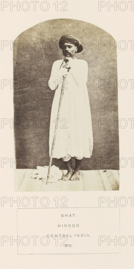 Portrait of an unknown man from the Bhat community with a rod, Madhya Pradesh, India, photograph, page, inscription: 'BHAT., HINDOO., CENTRAL INDIA.', number: '376.', photographer: James John Waterhouse (possibly), 1862, Central India, paper, albumen print, height 129 mm x width 79 mm, human races, peoples, nationalities, walking-stick, staff, A monochrome photograph of a barefoot man standing on a mat against a plain arched backdrop, wearing a long light-colored robe and a turban, holding a long staff vertically with both hands near his chest, with a folded cloth on the floor beside the staff and the man's feet visible at the hem of his robe.