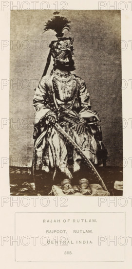 Portrat of Bharoo Singhji, the Raja of Ratlam, Madhya Pradesh, India, photograph, page, inscription: 'RAJAH OF RUTLAM., RAJPOOT. RUTLAM., CENTRAL INDIA.', number: '385.', photographer: James John Waterhouse (possibly), Ratlam, 1862, Central India, paper, albumen print, albumendruk, height 130 mm x width 80 mm, ruler, sovereign, human races, peoples, nationalities, A sepia-toned photograph of a seated man wearing ornate traditional clothing and layered jewelry, including necklaces and a decorated headpiece topped with a tall plume; he has facial hair and holds a long curved sword resting across his lap, with richly patterned fabric draped around his legs and visible footwear placed on a cushion; the background is plain and the image is framed by margins with a pale border.