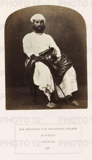 Portrait of Tukoji Rao Holkar, the Maharaja of Indore, Madhya Pradesh, India, photograph, page, inscription: 'HIS HIGHNESS THE MAHARAJAH HOLKAR., HINDOO., INDORE.', number: '387.', photographer: James John Waterhouse (possibly), Indore, 1862, Central India, paper, albumen print, height 122 mm x width 96 mm, ruler, sovereign, human races, peoples, nationalities, A sepia photograph of a seated man wearing a light turban and a light long-sleeved garment with layered cloth draped over his lap, a dark beaded necklace, and a beard; he sits on a chair with one hand resting on the draped fabric and the other holding a long slender staff that extends to the floor, his footwear is ornate and pointed; the background is a plain dark backdrop and the image has rounded top corners with a lighter border surrounding it.