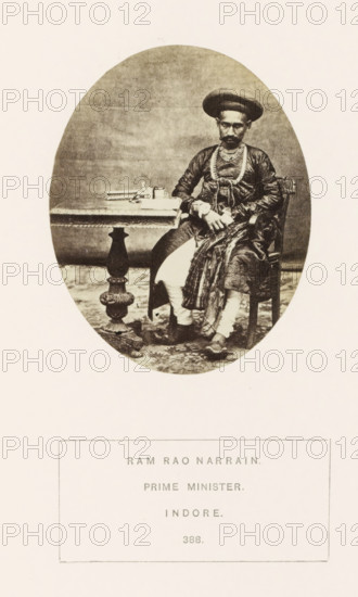 Portrait of Ram Rao Narrain, a prime minister of the princely state of Holkar, Indore, Madhya Pradesh, India, photograph, page, inscription: 'RAM RAO NARRAIN., PRIME MINISTER., INDORE.', number: '388.', photographer: James John Waterhouse (possibly), 1862, Central India, paper, albumen print, height 93 mm x width 75 mm, prime minister, human races, peoples, nationalities, An oval sepia photograph shows a seated man wearing a rounded turban and layered clothing, including a patterned shawl draped over one shoulder and decorative necklaces; he sits on a carved wooden chair with one hand resting on his lap and the other near a small table beside him, which holds stacked books and a small container; the table has an ornate pedestal base and the scene is set against a plain backdrop with a patterned floor covering visible beneath the chair and table.