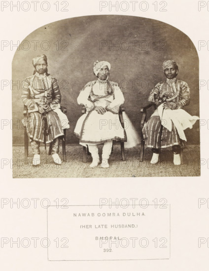 Portrait of Oomra Dulha, husband of Sekunder Begum, a queen of Bhopal, Madhya Pradesh, India, photograph, page, inscription: 'NAWAB OOMRA DULHA., (HER LATE HUSBAND.), BHOPAL.', number: '392.', photographer: James John Waterhouse (possibly), Bhopal, 1862, Central India, paper, albumen print, height 102 mm x width 118 mm, ruler, sovereign, human races, peoples, nationalities, A sepia-toned photograph shows three seated men wearing patterned robes and turbans, each sitting on a wooden chair against a plain backdrop; the central figure wears a lighter outfit with decorative trim and has hands folded in his lap, while the two flanking figures wear striped garments and hold canes or staffs with cloth draped over their arms, all three wearing footwear and positioned on a patterned floor covering.