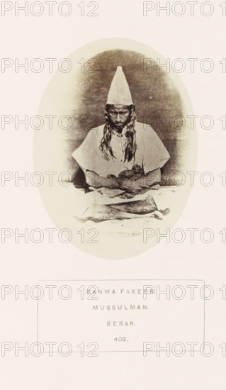 Portrait of an unknown fakir from the former Berar in central India, photograph, page, number: '402.', inscription: 'BANWA FAKEER., MUSSULMAN., BERAR.', photographer: James Mulheran (possibly), Berar, 1862, paper, albumen print, height 103 mm x width 77 mm, human races, peoples, nationalities, asceticism, Hinduism, Buddhism, Jainism, An oval sepia photograph shows a person seated facing forward wearing a tall conical hat and a loose, light-colored garment with short sleeves; the person's long hair falls over the shoulders and they look downward with hands resting in their lap holding a cloth or bundle, and a rectangular object lies on the surface in front of them, all set against a softly shaded background within the oval, surrounded by a pale border and further pale margins.
