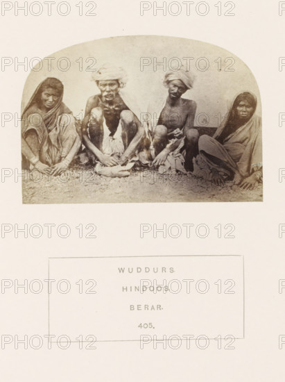 Portrait of four unknown people from the Waddar tribe of the former Berar in central India, photograph, page, inscription: 'WUDDURS., HINDOOS., BERAR.', number: '405.', photographer: James Mulheran (possibly), Berar, 1862, paper, albumen print, height 66 mm x width 95 mm, human races, peoples, nationalities, A faded oval photograph shows four seated people on a textured ground against a plain background, two men in the center wearing cloths wrapped around their heads and shoulders and two women at the sides draped in shawls, all sitting with legs drawn close and hands resting near their feet, wearing simple garments and appearing in sepia tones with visible grain and slight discoloration around the edges.