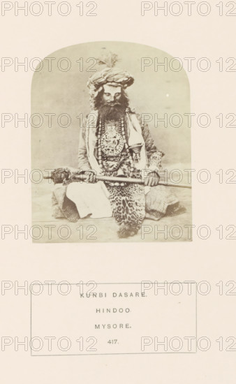 Portrait of an unknown man from the Kunbi community with ceremonial clothes, Mysore, Karnataka, India, photograph, page, inscription: 'KUNBI DASARE., HINDOO., MYSORE.', number: '417.', c. 1865 - in or before 1875, photographer, South India, paper, albumen print, height 95 mm x width 75 mm, head-gear: turban, human races, peoples, nationalities, ritual practices with particular objectives, Hinduism, Buddhism, Jainism, A sepia-toned photograph framed by a rounded rectangular border shows a seated man wearing a large wrapped turban with a plume, a full beard, and layered garments including a patterned robe and a draped cloth; he holds a long staff horizontally across his lap with both hands, and several beaded necklaces and a large ornate medallion hang over his chest, while a patterned textile is draped over his legs and he sits on a flat surface against a plain backdrop.