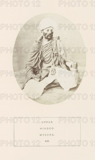 Portrait of an unknown man from the Uppara community of Mysore, Karnataka, India, photograph, page, number: '418.', inscription: 'UPPAR., HINDOO., MYSORE.', Mysore, c. 1865 - in or before 1875, photographer, South India, paper, albumen print, height 107 mm x width 88 mm, head-gear: turban, human races, peoples, nationalities, An oval sepia photograph shows a seated man wearing a turban and layered draped garments, with multiple long beaded necklaces and decorative cords around his neck and shoulders; he has a beard and moustache and is looking slightly to one side, one hand resting on his knee and the other holding a cylindrical object, while the background inside the oval is plain and the photograph is mounted on a light rectangular page.