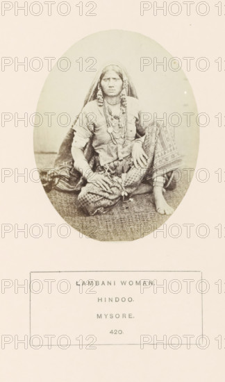 Portrait of an unknown woman from the Lambani tribe of Mysore, Karnataka, India, photograph, page, The Lambani tribe is also known as Banjara., number: '420.', inscription: 'LAMBANI WOMAN., HINDOO., MYSORE.', Mysore, c. 1865 - in or before 1875, photographer, South India, paper, albumen print, height 98 mm x width 76 mm, head-gear (with NAME), human races, peoples, nationalities, A sepia-toned oval photograph shows a seated woman wearing a head covering and layered necklaces, with patterned trousers and a sash tied at the waist; she sits barefoot on a textured mat with one knee raised and hands resting on her legs, her expression neutral and her hair partially visible beneath the veil, and the photograph is set on a pale background with a rectangular bordered area below.
