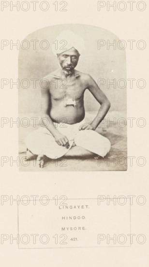 Portrait of an unknown man from the Lingayat community of Mysore, Karnataka, India, photograph, page, number: '421.', inscription: 'LINGAYET., HINDOO., MYSORE.', Mysore, c. 1865 - in or before 1875, photographer, South India, paper, albumen print, height 103 mm x width 76 mm, human races, peoples, nationalities, A sepia-toned portrait of a seated man framed within an arched vignette, showing a bare-chested figure wearing a light turban and a light lower garment, seated cross-legged on the ground with one hand resting on his knee and the other hand near his lap, a small pendant or object suspended on a cord around his neck, a neutral plain background behind him, and a rectangular bordered area below the portrait containing printed lines.
