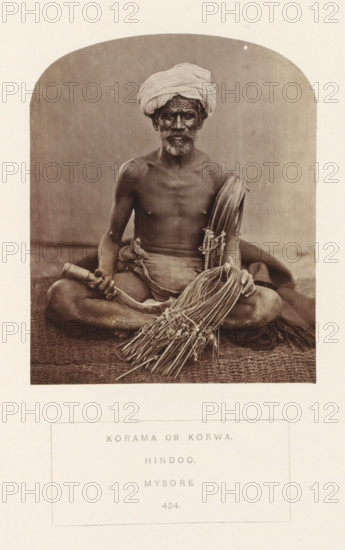 Portrait of an unknown man from the Korama community with a rope used to make a basket or a mat, Mysore, Karnataka, India, photograph, page, number: '424.', inscription: 'KORAMA OR KORWA., HINDOO., MYSORE.', c. 1865 - in or before 1875, photographer, South India, paper, albumen print, height 115 mm x width 94 mm, human races, peoples, nationalities, handicrafts and industries, A sepia-toned photograph of a seated man on a woven mat, shown from head to thighs within an arched frame; he wears a wrapped turban and has a beard and mustache, his torso is bare and he is draped in cloth around his waist; he sits cross-legged holding a curved wooden handle in one hand and a bundled collection of long thin reeds or cords in the other, with additional tied bundles resting across his lap and beside him; the background behind him is plain and softly textured.