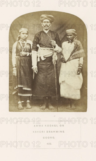 Portrait of three people from the Kodava community of Kodagu. Karnataka, India, photograph, page, number: '425.', inscription: 'AMMA KODAGI, OR, KAVERI BRAHMINS., COORG.', photographer: G. Richter (possibly), Kodagu, c. 1865 - in or before 1875, South India, Karnataka, paper, albumen print, height 122 mm x width 95 mm, human races, peoples, nationalities, A sepia photograph shows three barefoot people standing side by side against a plain backdrop, with a man in the center wearing a dark long garment, a wrapped sash at his waist, a turban, and holding a long staff while a curved knife rests at his side; a girl on his left wears a dress with decorative trim, multiple necklaces, bracelets, and anklets, and has a cloth tied around her head; a person on his right wears a light wrapped garment with a shawl draped over one shoulder and a head covering, and also wears bracelets.