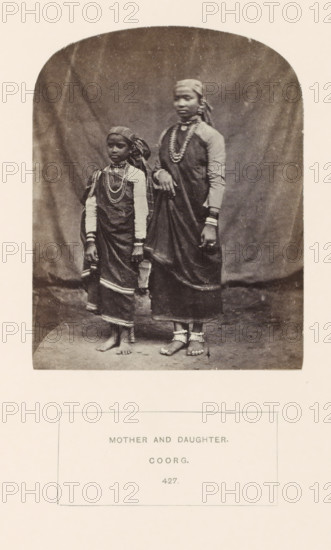 Portrait of an unknown mother and daughter from the Kodava community of Kodagu, Karnataka, India, photograph, page, number: '427.', inscription: 'MOTHER AND DAUGHTER., COORG.', photographer: G. Richter (possibly), Kodagu, c. 1865 - in or before 1875, South India, paper, albumen print, height 123 mm x width 97 mm, mother and daughter(s) (family group), human races, peoples, nationalities, A sepia-toned photograph shows two females standing on a plain floor against a draped fabric backdrop; both wear long dark garments with lighter long-sleeved underlayers and patterned headscarves, and both are adorned with multiple layered necklaces and bracelets; the taller figure rests one hand on the shorter figure’s shoulder and wears anklets and toe rings while the shorter figure’s bare feet are visible and she has a cloth draped over one arm; the image has rounded top corners and is mounted on a lighter paper.