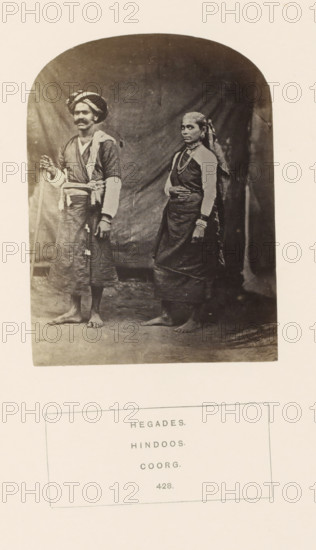 Portrait of Meder and his wife, cultivators from the Hegde community of Kodagu, Karnataka, India, photograph, page, number: '428.', inscription: 'HEGADES., HINDOOS., COORG.', photographer: G. Richter (possibly), Kodagu, c. 1865 - in or before 1875, South India, paper, albumen print, height 116 mm x width 87 mm, human races, peoples, nationalities, A sepia photograph shows a man and a woman standing barefoot on a plain surface against a draped backdrop, the man on the left wearing a turban, a layered garment with a sash and loose lower wrap, holding a staff, and the woman on the right wearing a draped sari-like garment with multiple necklaces, bangles, and a headscarf, both facing forward with neutral expressions.