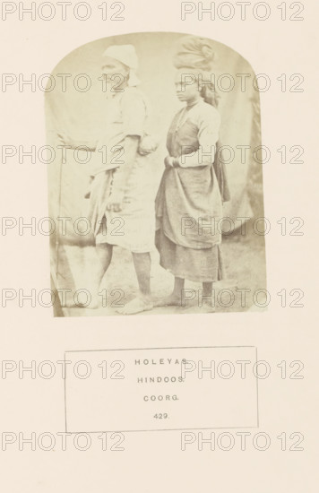 Portrait of an unknown man and woman from the Holeya community of Kodagu, Karnataka, India, photograph, page, inscription: 'HOLEYAS., HINDOOS., COORG.', number: '429.', photographer: G. Richter (possibly), Kodagu, c. 1865 - in or before 1875, South India, paper, albumen print, height 115 mm x width 88 mm, human races, peoples, nationalities, A faded sepia photograph shows two barefoot individuals standing side by side against a plain backdrop, the person on the left facing sideways and holding a long, thin staff while wearing a short-sleeved garment with a wrapped lower cloth and a head covering, and the person on the right standing slightly behind with hands clasped in front, wearing layered garments including a sleeveless outer piece over a long-sleeved top and a wrapped lower skirt, with a cloth wrapped around their head and hair visible at the back.