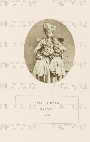 Portrait of an unknown boy from the military class in Mysore, Karnataka, India, photograph, page, inscription: 'RAJAH BINDAH., MYSORE.', number: '408.', Mysore, c. 1865 - in or before 1875, photographer, South India, paper, albumen print, height 79 mm x width 63 mm, human races, peoples, nationalities, An oval sepia photograph of a seated young person wearing a patterned turban and layered traditional clothing, including a draped patterned shawl over one shoulder and beaded necklaces, sitting on a wooden chair with arms and looking toward the camera, placed above a rectangular caption box on a pale background.