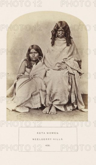 Portrait of two unknown women from the Kota tribe of the Nilgiri mountains in Tamil Nadu, India, photograph, page, inscription: 'KOTA WOMEN., NEELGERRY HILLS.', number: '436.', Nilgiris, c. 1865 - in or before 1875, photographer, South India, Tamil Nadu, paper, albumen print, height 145 mm x width 107 mm, human races, peoples, nationalities, A sepia-toned photograph shows two seated women wrapped in plain draped cloths, one sitting slightly lower and leaning against the other, both with long dark hair and large circular earrings; the taller woman has her hands folded over her lap and bare feet visible at the bottom edge of the drapery, while the shorter woman wears multiple necklaces and bracelets and looks slightly away from the camera; the background is a plain neutral backdrop and the image is bordered by a rounded top frame.