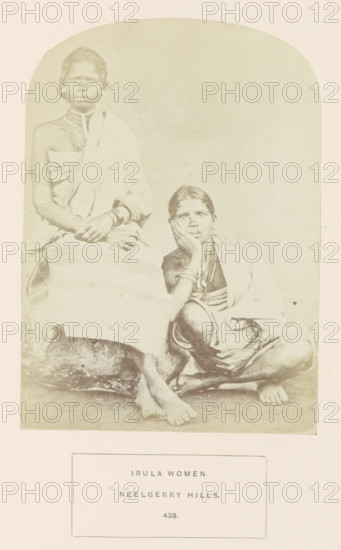 Portrait of two unknown women from the Irula tribe of the Nilgiri mountains in Tamil Nadu, India, photograph, page, number: '438.', inscription: 'IRULA WOMEN., NEELGERRY HILLS.', Nilgiris, c. 1865 - in or before 1875, photographer, South India, Tamil Nadu, paper, albumen print, height 165 mm x width 119 mm, human races, peoples, nationalities, A faded sepia photograph shows two seated women wearing draped cloth garments and multiple necklaces and bracelets; one woman sits upright with her hands resting in her lap while the other sits lower with an elbow propped on a knee and her hand supporting her face, both barefoot on a textured ground, with a plain light background and a rectangular bordered panel beneath the image.