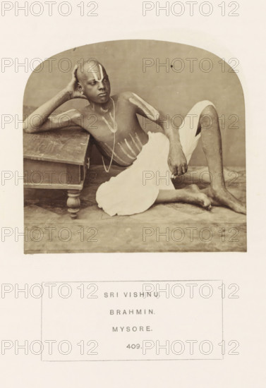 Portrait of an unknown priest from the Vaishnav Brahmin community of Mysore, Karnataka, India, photograph, page, number: '409.', inscription: 'SRI VISHNU., BRAHMIN., MYSORE.', Mysore, c. 1865 - in or before 1875, photographer, South India, paper, albumen print, height 91 mm x width 95 mm, Brahma (supreme being, creator, etc., Hinduism, Buddhism, Jainism), human races, peoples, nationalities, A sepia photograph shows a seated man reclining beside a low wooden table, his torso bare and a light cloth wrapped around his lower body, with his left arm resting on the table and his right hand placed on his knee; his head is shaved and marked with light painted lines on the forehead, chest, shoulders, and arms, and he wears a thin necklace, while the background is plain and the floor beneath him appears textured.