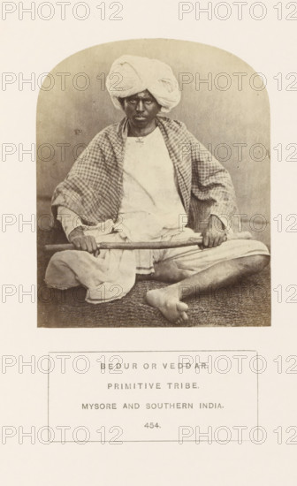 Portrait of an unknown man with a stick in his hands, Mysore, Karnataka, India, photograph, page, number: '454.', inscription: 'BEDUR OR VEDDAR., PRIMITIVE TRIBE., MYSORE AND SOUTHERN INDIA.', c. 1865 - in or before 1875, photographer, South India, paper, albumen print, height 107 mm x width 86 mm, head-gear: turban, human races, peoples, nationalities, A seated person wearing a wrapped turban and a light garment with a checked shawl draped over the shoulders is shown in a framed oval photograph; the person holds a long stick across their lap with both hands and sits cross-legged on a textured mat, with one bare foot visible, and a neutral backdrop behind them.